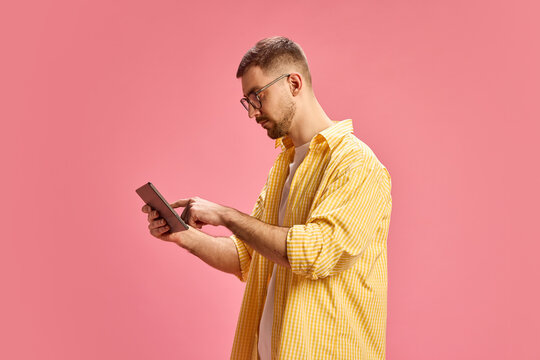Young man in yellow checkered shirt using digital tablet, standing against pink background. Concept of technology, communication, lifestyle, and modern business, online communication