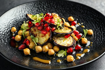 A top view of chickpea salad on a black plate background. Roasted eggplant arcs, vibrant pomegranate seeds, golden chickpeas, and lemon.