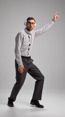 Full-length image of joyful young man in glasses and headphones, wearing shirt and dark jeans, dancing against grey studio background. Concept of human emotions, lifestyle, business, gadgets