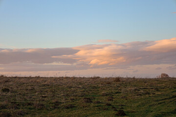 A serene scene of a rural landscape at sunset. Soft evening light illuminates a rolling terrain covered in grass and small bushes.