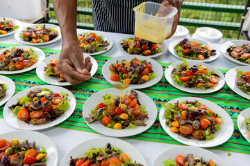 Close-up of a chef's hand pouring a creamy dressing over a fresh salad. The salad contains cherry...