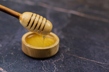 Close-up of yellow liquid honey dripping from a wooden honey spoon into a wooden bowl on a dark...