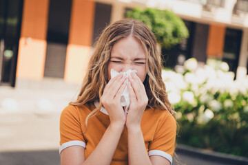Young woman outdoors, sneezing into tissue due to allergies during a sunny urban day, highlighting...