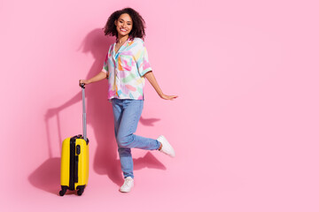 Young woman with curly hair smiling while posing with a yellow suitcase against a pink background,...