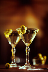 A martini glass with olives on a pick, placed in a warm, dimly lit setting with soft golden tones.