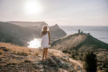 Naklejka premium A blonde woman stands on a hill overlooking the ocean. She is wearing a white dress and she is enjoying the view.