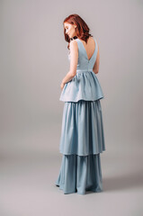 Fototapeta premium Sensual redhead woman in blue evening dress. Bridesmaid's fashion. Plunge neck sleeveless maxi gown with tiered skirt. Trendy outlook for hen party. Summer feminine waisted look, studio portrait, rear