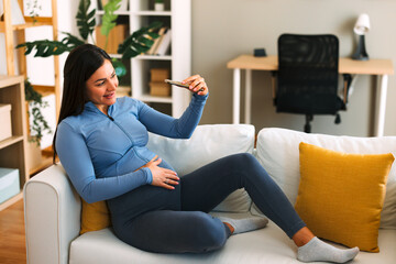 Obraz premium Happy pregnant woman taking a selfie with smartphone while sitting on a comfortable sofa at home