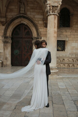 A romantic wedding moment in Croatia. The groom embraces the bride near an ancient building as her...