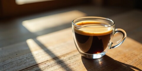 A single serving of dark roasted coffee sits in a clear glass cup, illuminated by sunlight streaming through a nearby window, casting shadows on a rustic wooden surface.