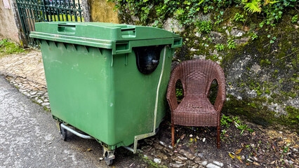 Green dumpster beside a brown wicker chair on a cobblestone street, urban waste management concept