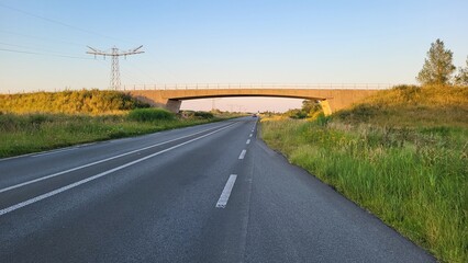 A concrete bridge spans across an empty highway surrounded by lush green grass. The clear sky reflects a warm glow as the sun sets in the background, creating a peaceful atmosphere.