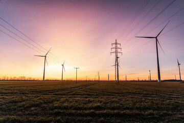 Wind turbines and power pylons with electrical lines are silhouetted against a vibrant, colorful sky during the golden hour, highlighting the harmony between renewable and traditional energy sources.
