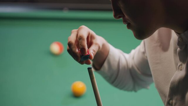 Young man in white shirt chalking cue stick before taking shot on green pool table. Close-up highlights focus, precision, and preparation, with billiard balls visible in background