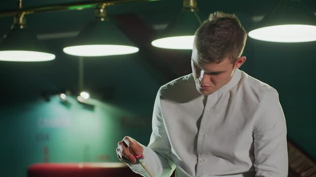 Close-up shot of chap in white shirt holding cue stick with serious face in dimly lit billiards hall. Overhead lighting highlights focused expression and intense atmosphere of professional gameplay