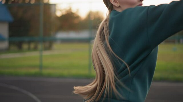 Woman in green hoodie serving volleyball, flapping hand as ball rises, with open field and building in background, action shot of athlete training outdoors