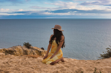 Young, beautiful brunette woman with straw hat is kneeling on the sand on the cliff. The girl is relaxed and looks at the horizon where the sky meets the sea. © @skuder_photographer