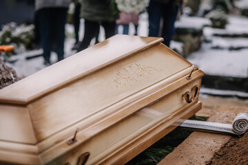 Close-up of a wooden coffin being lowered into a grave during a winter funeral, with mourners...