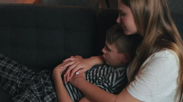 Compassionate sister providing emotional support, consoling her upset younger brother on a cozy sofa in a dimly lit room, embodying sibling bonding and empathy