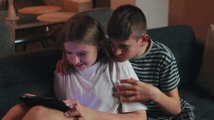 Young brother and sister sitting side by side on living room couch, engaging with digital tablet, showing wide-eyed wonder while browsing interactive screen
