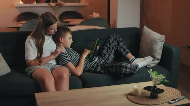 Teenage siblings sharing leisure moment, sister warmly hugging brother while playing videogame on tablet during relaxed evening at home living room