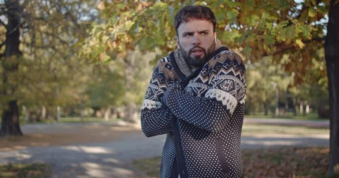 A man with a beard, bundled in a sweater, shivers in a chilly autumn park. His facial expressions convey feelings of coldness and discomfort.
