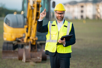 Construction owner near excavator. Confident construction owner in front of house. Architect, civil engineer. Man construction owner with a safety vest and hardhat at construction site.