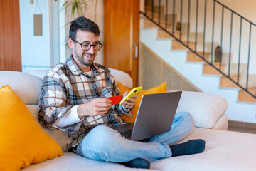 Smiling Man Shopping Online with Credit Card at Home. Happy man sitting on couch using smartphone and credit card for online shopping with laptop nearby.