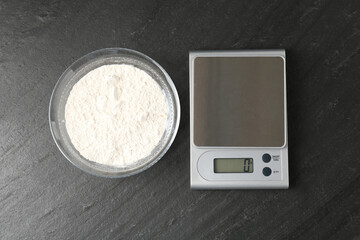Electronic kitchen scale and bowl with flour on dark textured table, flat lay