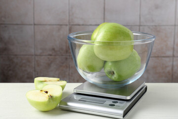 Electronic kitchen scale with bowl of green apples on white wooden table against tiled wall, closeup