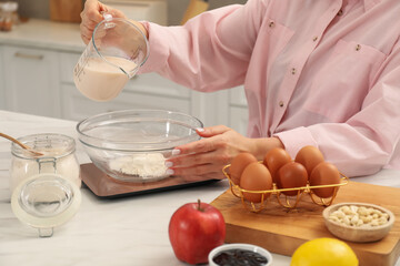 Woman pouring milk into bowl on electronic kitchen scale at white marble table indoors, closeup