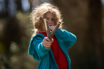 Portrait of a child shooter aiming. Funny childhood. Kids game with a slingshot. Kids handmade slingshot weapon. Kids toy sling weapon in hand. Shooting kids game. Kid attacking with a catapult.