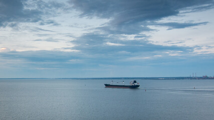 Naklejka premium Cargo ship sailing in the bay. Seascape, view from the high shore. Beautiful evening sky with clouds.