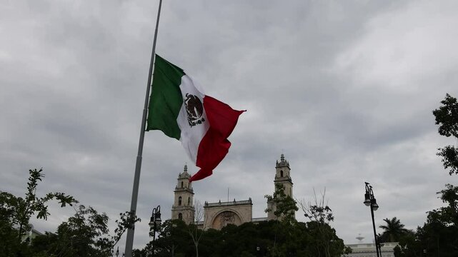 mexican flag waving in front of the cathedral in merida yucatan mexico on a cloudy overcast day in latin america (beautiful colonial spanish style architecture with mayan stones)