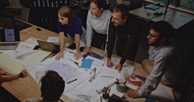 A diverse team of business colleagues collaborates on a project, reviewing blueprints and engaging in productive discussion around a table.