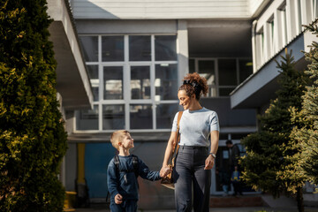 Mother and her kindergartener holding hands in front of school building while smiling at each other.