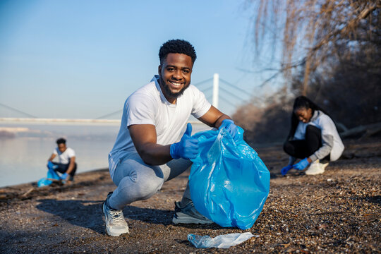 Eco conscious multicultural volunteer with garbage bag in hand cleaning riverbank from plastic waste with his friends and giving thumbs up at camera.