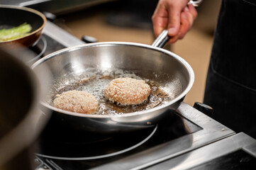 Close-up of hands frying breaded patties in a stainless steel pan, sizzling in oil, capturing a professional cooking process in a kitchen.