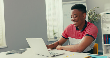 Trainee in office sits at desk in front of laptop monitor in background whiteboard, young dark-skinned man talks online with clients, co-workers, uses webcam, video call, smiling, laugh, waving hand