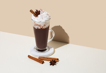 Hot chocolate with whipped cream, cinnamon and spices in a tall glass on a white and beige background
