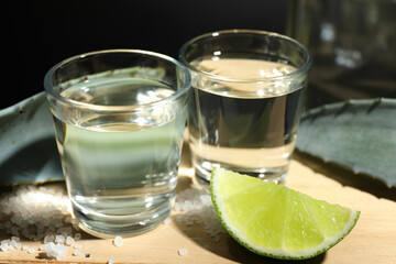 Tequila shots, slice of lime, salt and agave leaves on wooden table against black background, closeup