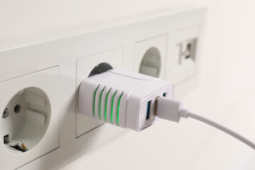 USB adapter with cable plugged into electric socket on light wall, closeup