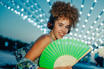 Young woman listening music with headphones and cooling with hand fan at summer festival