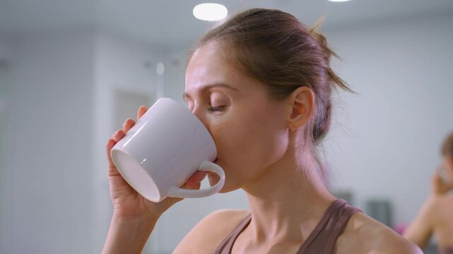 Woman with brown signet hair tied back drinks from coffee cup while mirror reflects her back and background, relaxing post-workout moment in gym setting