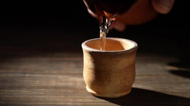 Japanese sake from a bottle being poured into ceramic cup on a wooden table, close up, slow motion