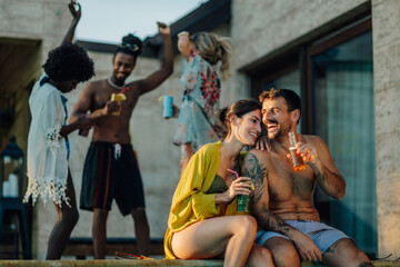 Happy couple enjoying drinks by the pool while friends dance in the background