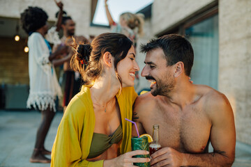 Happy couple enjoying drinks at a pool party with friends dancing in the background