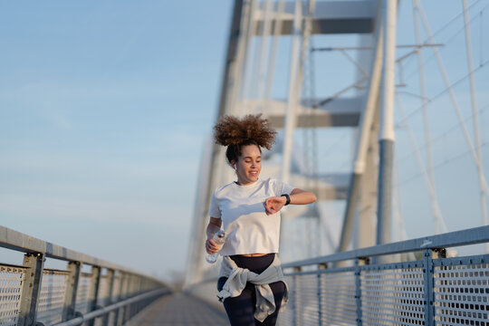 A young woman runs on a modern bridge while checking her smartwatch. She wears a white t-shirt, leggings, and a tied jacket, holding a water bottle. The clear sky enhances the urban fitness scene.