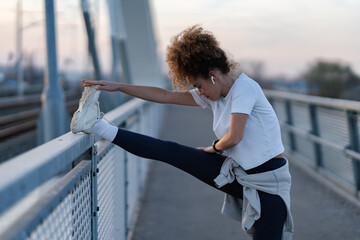Young woman with curly hair performs a deep leg stretch on a bridge railing during an outdoor...