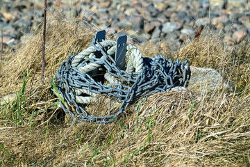 A coil of rusty chain and thick rope lies on dry grass and scattered rocks, illuminated by sunlight...